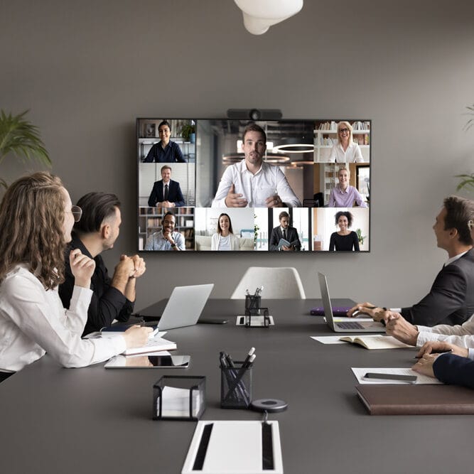 Alt-Text: Team in Video-Konferenz mit mehreren Teilnehmern auf einem großen Bildschirm, moderne Büroumgebung.